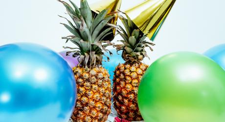 Two pineapple surrounded by balloons and party hats
