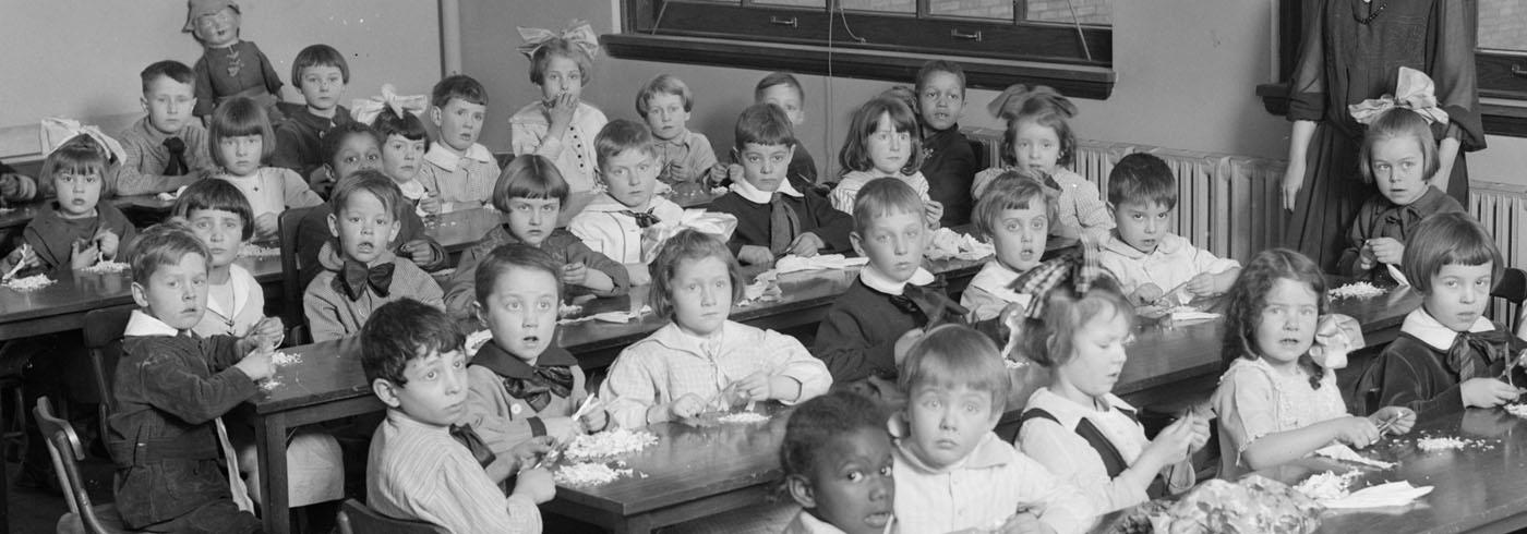 Victorian school kids, some with hair-bows as big as their heads, and lots of frills on their clothes. All crammed into a class room, cutting pieces of paper at the same time as looking bemused by the camera in their classroom