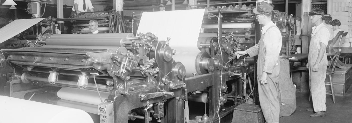 Several men stood around a newspaper printing press, with a few women sat on chairs behind them checking things.
