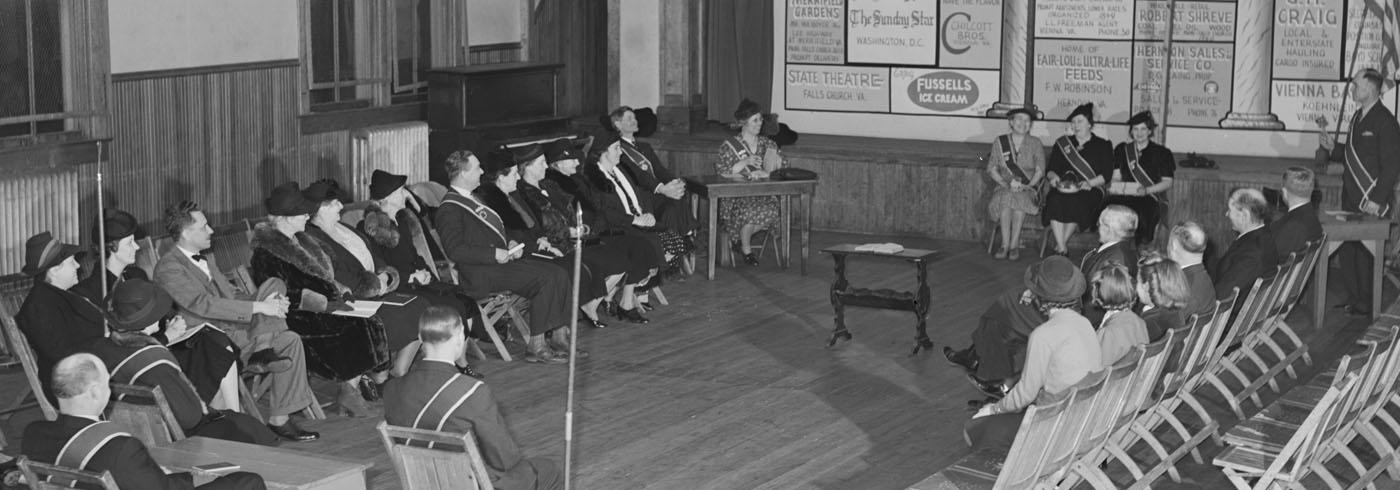 Smartly dressed people sat on fold-out chairs, in a rows facing each other in a timber clad town hall, with three women at the front looking very happy. Some empty chairs in the audience, like they were expecting a bigger turnout.