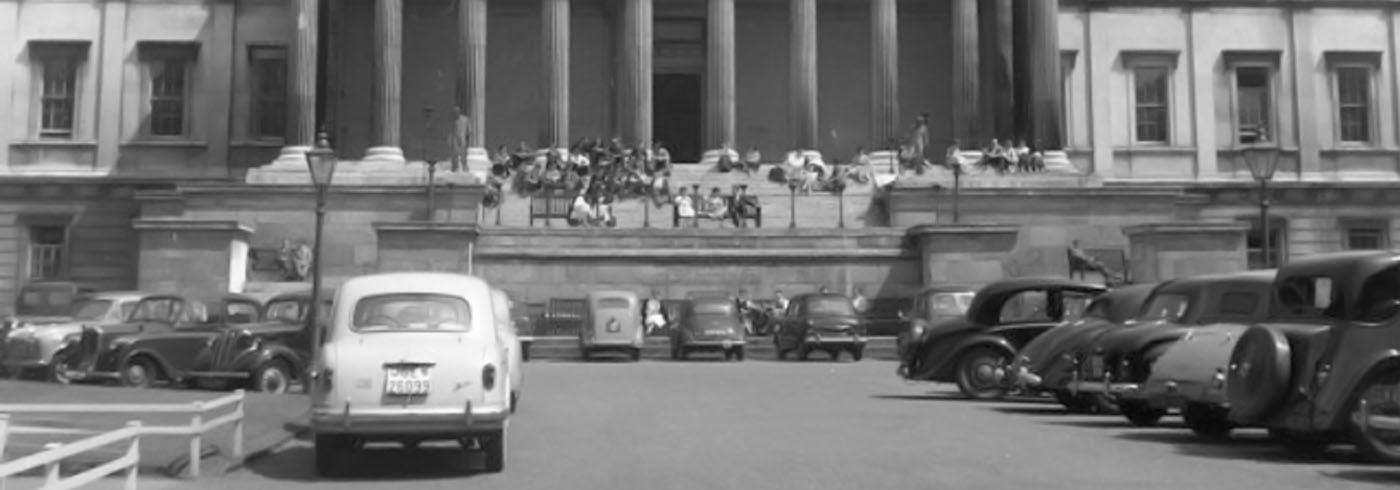 Students eating on the steps of an old stone building, with old cars parked in the carpark in the foreground.