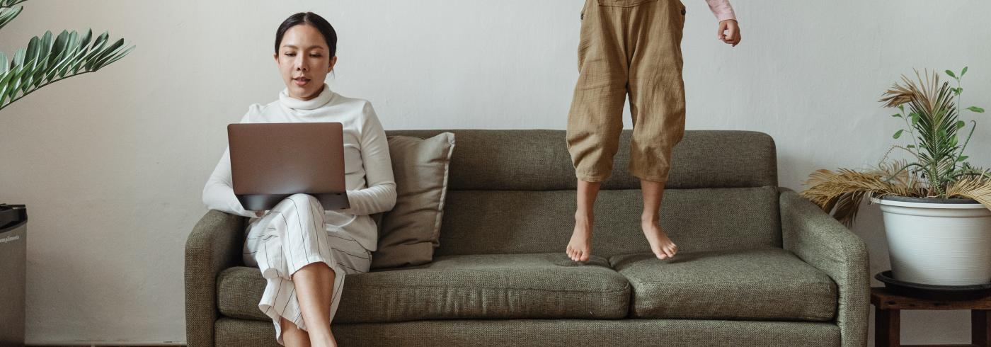 A woman working on her sofa and her child is next to her, jumping on the sofa