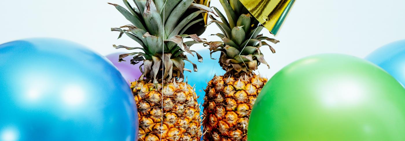 Two pineapple surrounded by balloons and party hats