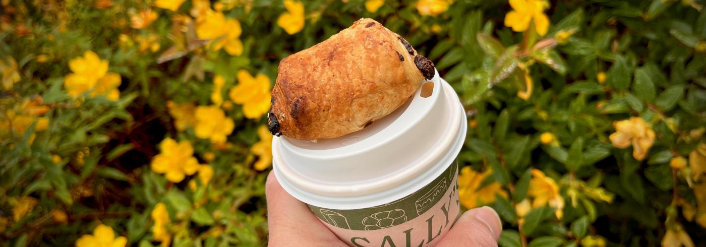 A hand holding a take away coffee cup with a croissant balanced on top with yellow flowers in the background.