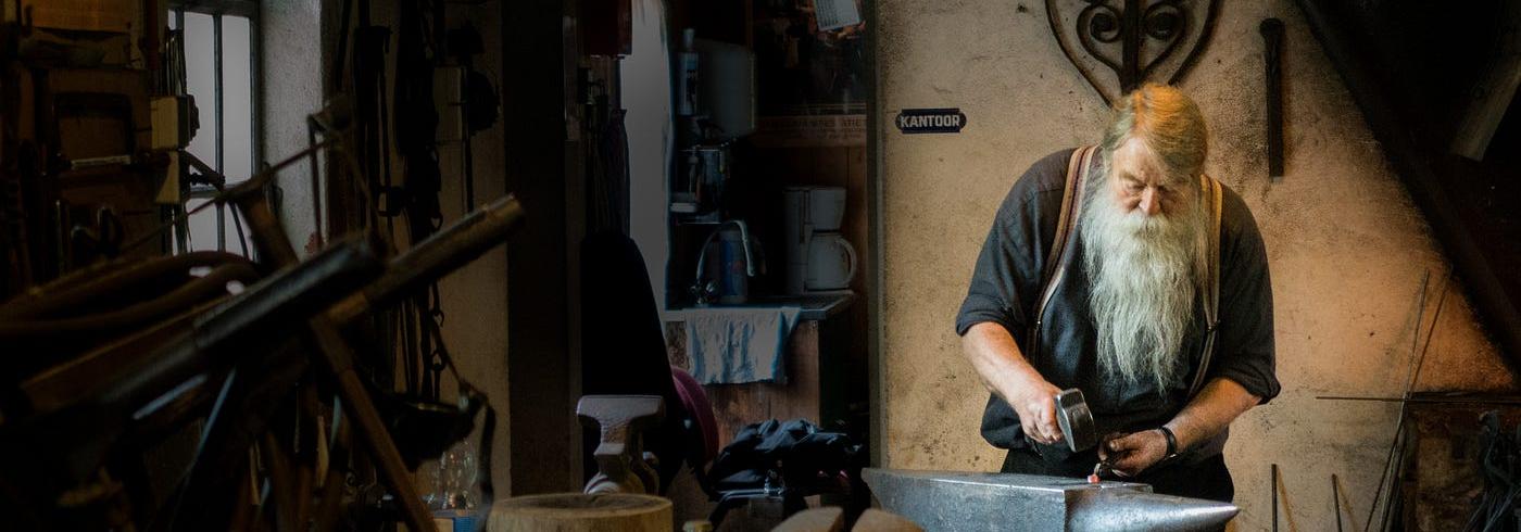 An old man in a workshop carefully crafting a piece of metal with a hammer and an anvil.